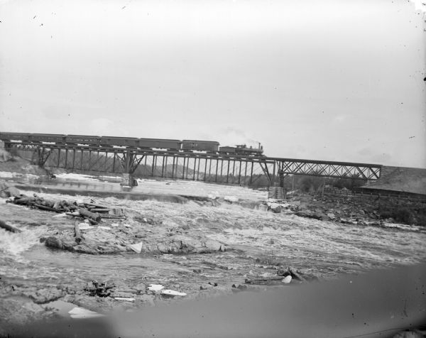 View across river towards a railroad train on a railroad bridge crossing the river. There are signs and graffiti painted on the rocks below.