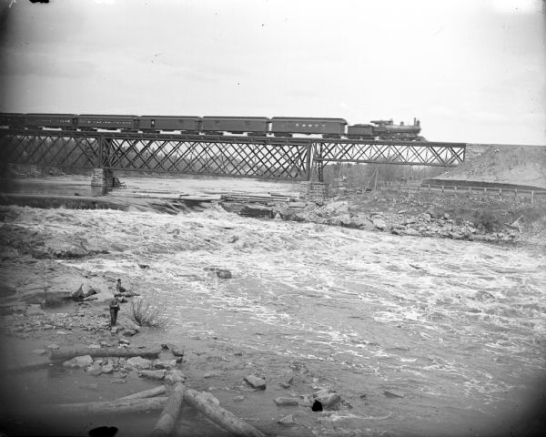 Elevated view looking across river of a railroad train on a railroad bridge crossing a river. On the rocks in the foreground below is a man fishing with a long fishing pole. Another person sits on a rock behind him.	