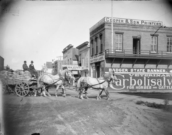View down street towards two men riding in a loaded wagon pulled by a team of four horses in front of the Palace Bakery and the offices for Cooper & Son Printers.