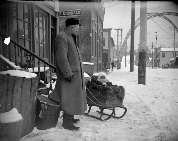 Winter scene with a man standing in a coat next to a child bundled in a sled on the snow-covered sidewalk in front of the storefront of J.R. McDonald. In the background is an arch over an intersection.	
