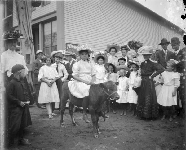 A crowd standing in a circle is watching a girl sitting on a calf. In the background is a storefront.