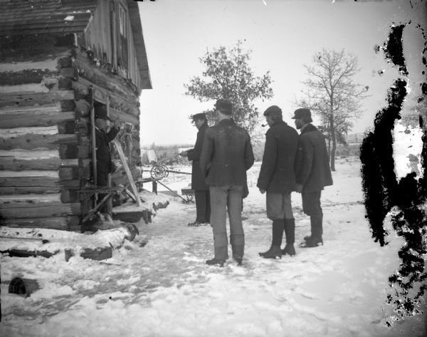 Group of men standing in the snow in front of a log house. One man is armed with a shotgun, facing a man standing in the open doorway of a log house.
