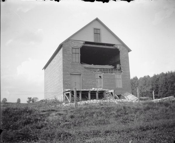 View up hill towards a large brick building with a large hole blasted in the second-floor. A wood platform below is collapsed.