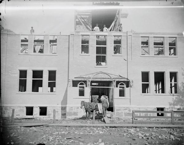 View towards front entrance of the partially constructed Senior High School.	A man is walking up a board into the open doorway of the school. A horse is standing nearby. Other workers are standing above the second story on platforms. The school has no roof.