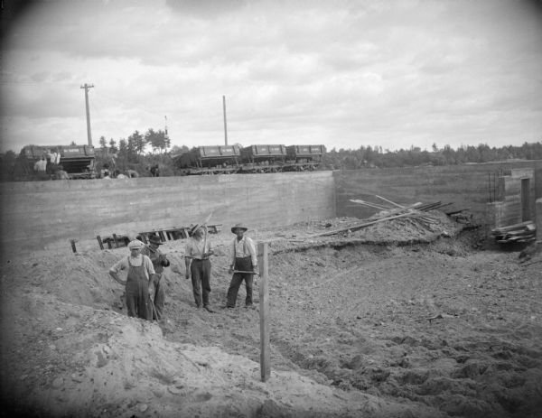 Four men in the foreground are posing standing with shovels in a large hole in the ground. In the background behind a wall, men are working near cars along railroad tracks near a man wearing a suit.