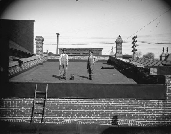 View from rooftop of two men posing standing on top of the roof of a brick building. There is a ladder on the left leading to the roof. The men are holding tools, and a hammer is lying on the roof. Roofs and chimneys are on the left and right, and a power line is in the far background.	