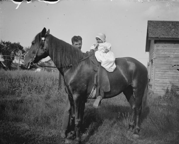 Man holding a baby sitting on a saddled horse.	