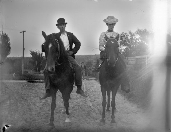 Man and woman posing side-by-side on horseback on a rutted road. The woman is riding sidesaddle. Another man is standing in the far background.