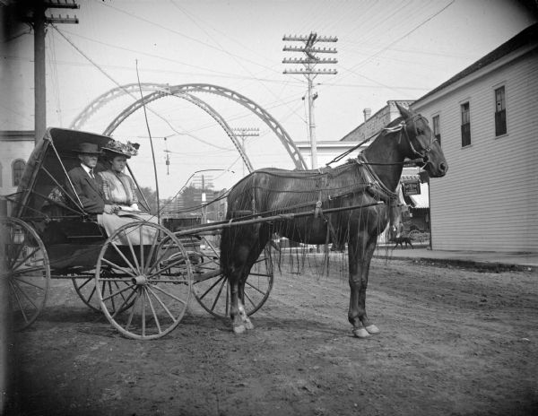 View down unpaved First Street towards a man and woman posing sitting in a buggy, pulled by a single horse wearing a fly-net. Behind them is an arch over the intersection flanked by large power poles. On the right a dog is on the sidewalk on the corner.