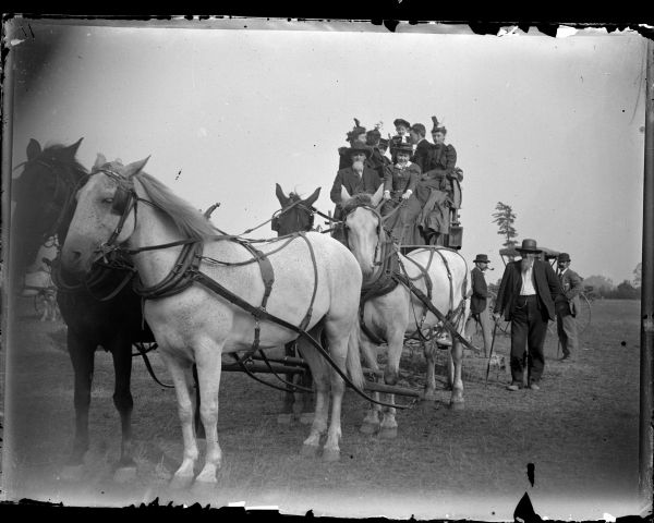 View from front of a group of people sitting on top of a stagecoach pulled by a team of four horses. Three men are standing on the right, with a horse-drawn buggy parked behind them.
