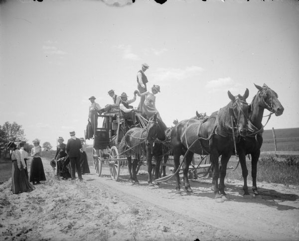 View from side of road towards men and women disembarking from a stagecoach pulled by a team of four horses. Two of the horses are wearing fly-nets.