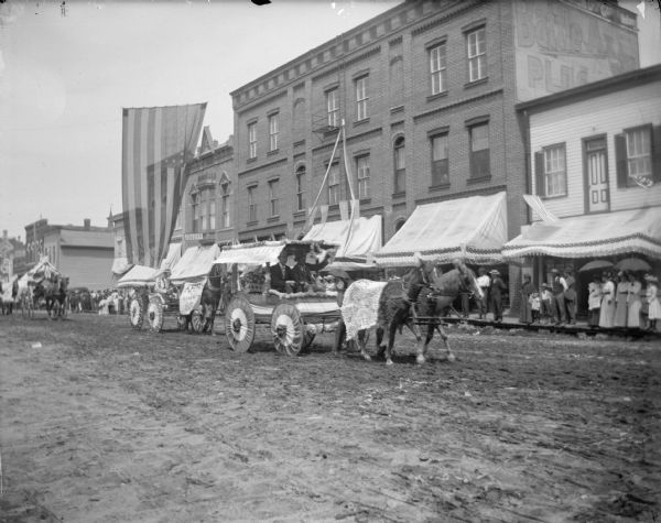 View across unpaved Main Street towards a crowd gathered to watch a parade of wagons.	