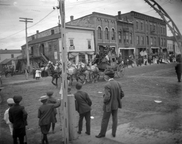 View from sidewalk of crowd gathered on sidewalk along Main Street to watch a patriotically decorated stagecoach pulled by a team of four horses passing by in a parade. In the foreground a man and a number of boys are standing on the sidewalk.	