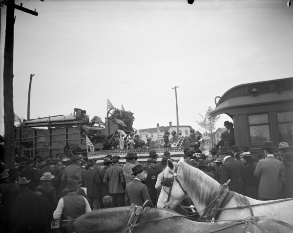 Elevated view over horses towards people gathered around a band on a platform decorated with flags. In the background on the left near the band is a large piece of equipment, perhaps a combine, decorated with flags. On the right men are standing on the end of a caboose.
