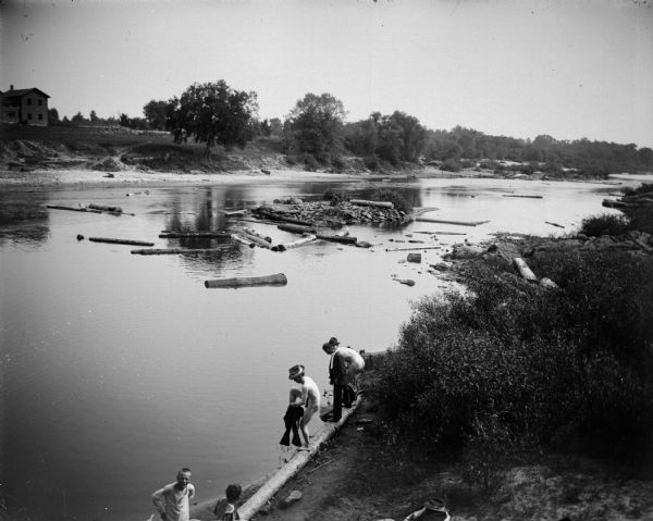 Slightly elevated view looking down at two men undressing to go skinny dipping in the river. A man wearing a suit is standing smiling between them, and two children are in the foreground. There are logs floating in the river, and a house on the far shoreline.