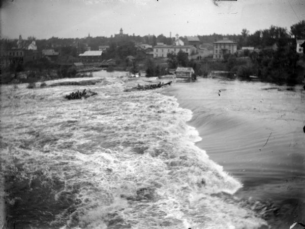 Elevated view across river at flood stage, with piles of logs and timber caught near rocks. A flooded small building is surrounded by water, and has an advertisement for "salve" painted on the sides and roof. On the far shoreline is the town of Black River Falls.