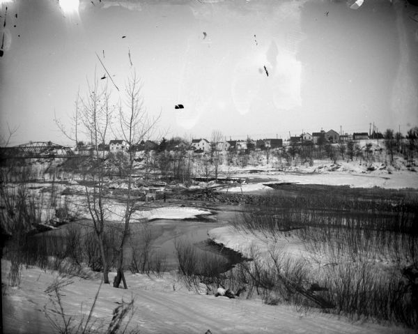 View from shoreline over river, with the buildings of town and a bridge on the opposite shoreline.	