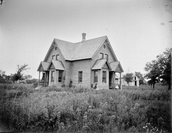 View across yard with tall plants towards three children sitting on one of the porches of a brick house. There is another porch on the left side of the house.