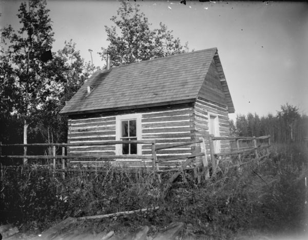 One room wooden cabin with a shingle roof surrounded by a post fence.