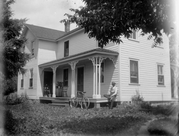 View across yard towards a man posing sitting on the porch of a frame house. There is a bicycle leaning against the porch.