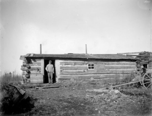 Man posing standing in the doorway of a low log cabin.	