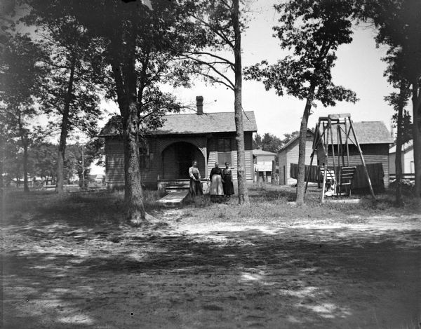 View across yard towards three women posing standing in the yard in front of a frame house near a wooden walkway. On the right a young girl is sitting nearby on a lawn swing.