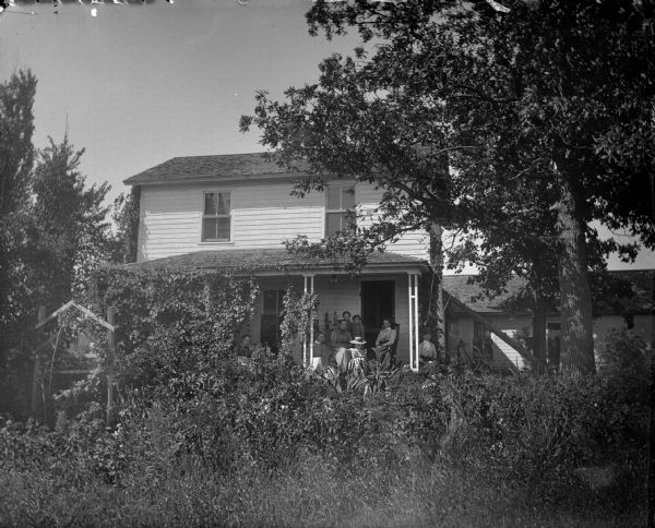 View across yard, with thick foliage and trees, towards a group of women and girls posing on the porch of a frame house. The porch is partially covered by vines.