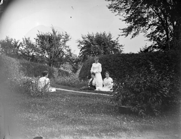 View across Spaulding House grounds towards four women posing. A woman on the left is sitting on the ground near bushes, and across a path two other women are also sitting on the ground while one woman is standing and placing what may be a flower in the hair of one of the women.