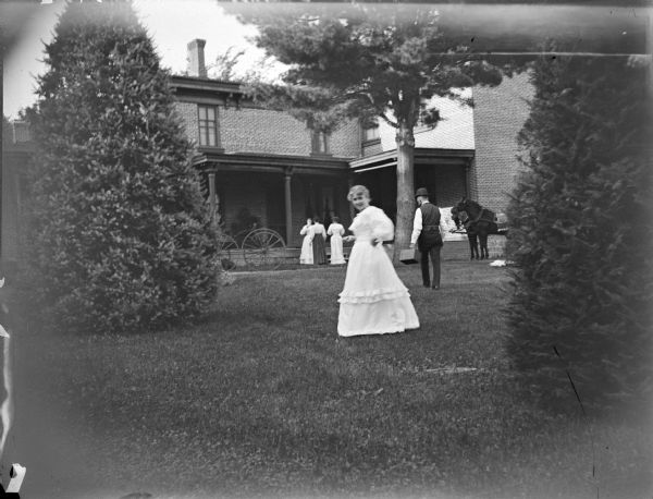 View across lawn towards four women and a man walking on the grounds in front of the Spaulding House. On the right near the house are two horses, and on the left in front of the porch is a carriage.