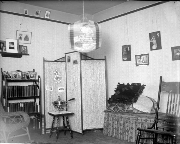 Interior of a furnished bedroom, probably a girl's room. A folding room screen is in the corner in the center, with a bookcase on the left. Framed images are hanging from picture rail molding along the wall near the ceiling.