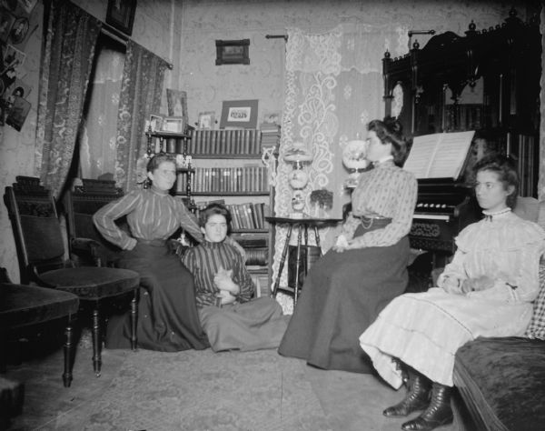 Four women are posing sitting in a parlor in front of an organ. One of the women is sitting on the floor holding a kitten.