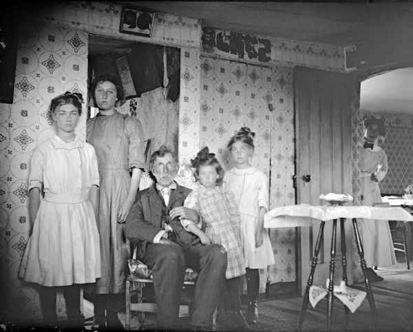 In the center is a man posing sitting in a chair. Beside him on the left are two teenage girls standing near a doorway, and on the right standing next to him are two younger girls. On the far right another young girl or woman is looking into the room from a doorway.