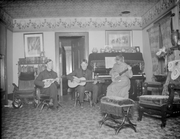 Two men and a woman are posing sitting in a parlor and holding musical instruments. The woman is sitting on the right in front of a piano playing a banjo. The two men are sitting in the center in front of a doorway. One is playing a mandolin, and the other man is playing a guitar.