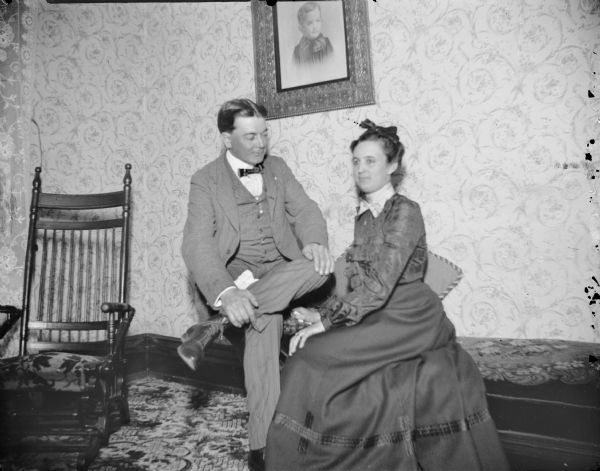 Man and woman posing sitting on a settee near the corner of a room.