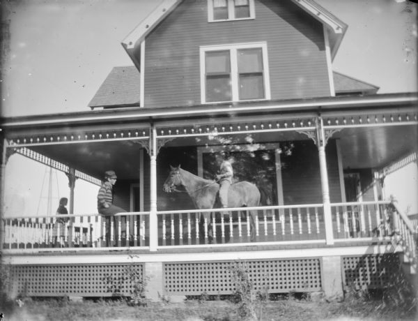 View from yard towards a boy posing sitting bareback on a horse on the wrap-around porch of a frame house. Another boy is sitting on the railing on the left nearby, and a girl is standing on the porch behind him in the background.	