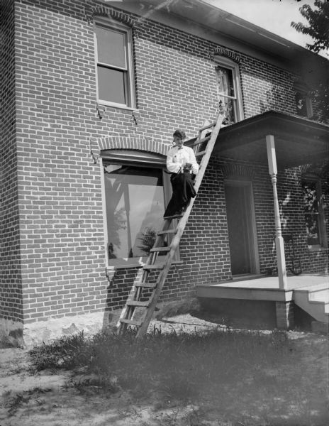 View across yard towards a woman posing sitting halfway up a ladder which is against the porch roof of a brick building. She is holding a kitten on her knee.
