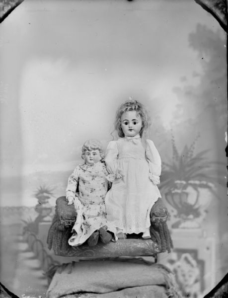 Studio portrait in front of a painted backdrop of two dolls propped up in a chair on a stool.