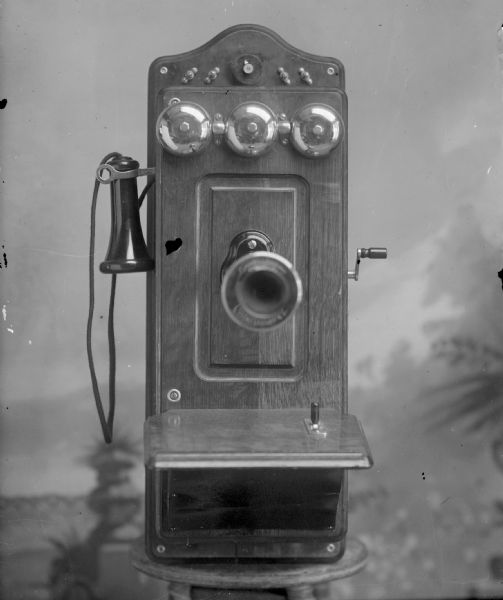 Kellogg box telephone, patented November 26, 1901, set on a stool in front of a painted background.	