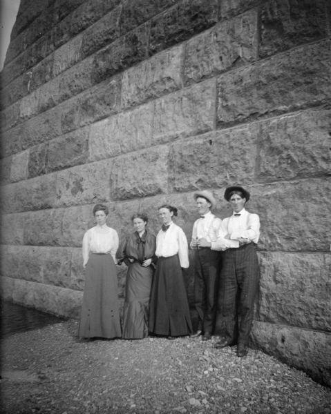 Three women and two men posing standing on gravel at the base of a stone wall. To the left is water.