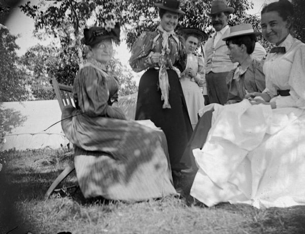 Three women are posing sitting, and two women and a man are standing under the shade of a tree. The woman in the center is holding a fish on a stringer. In the background is a tent.	