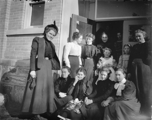 Group portrait of ten young women and two girls posing in the partially open doorway on the stoop of a brick building, probably the Union High School.
