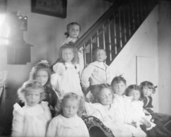 Group portrait of ten young children. Some of the children are posing standing on and around a staircase, and the others are sitting and standing on a sofa. There is a telephone on the wall on the left.