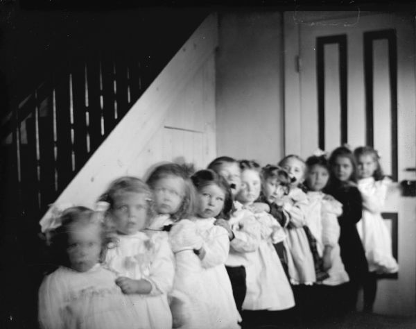 Group portrait of eleven children standing in a line along the side of a staircase. There is a closed door in the background.
