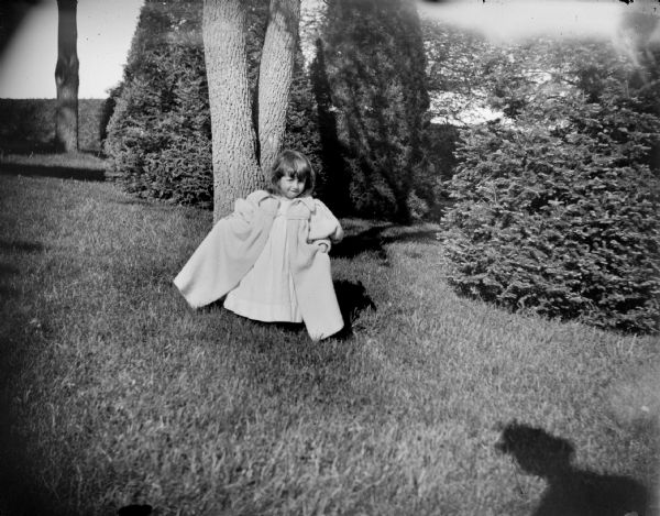 A young girl wearing a long coat is posing standing on the grounds of the Spaulding House. The shadow of a man wearing a hat is on the lawn the right foreground.