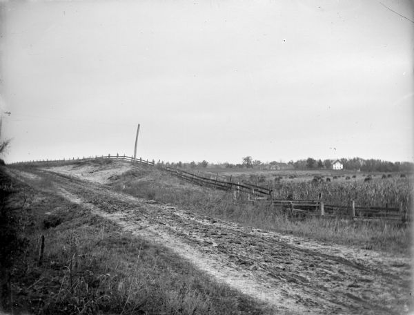 Unpaved road on a slope bordered by a fenced hay field. A house and outbuildings are in the far background.