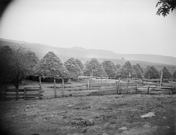 Haystacks in a field behind a wooden fence. Hills and bluffs are in the far background.
