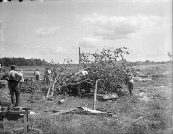 Men and women examining debris in a field, possibly caused by a tornado. A tree is on top of an automobile in the center. In the far distance are fields and buildings.