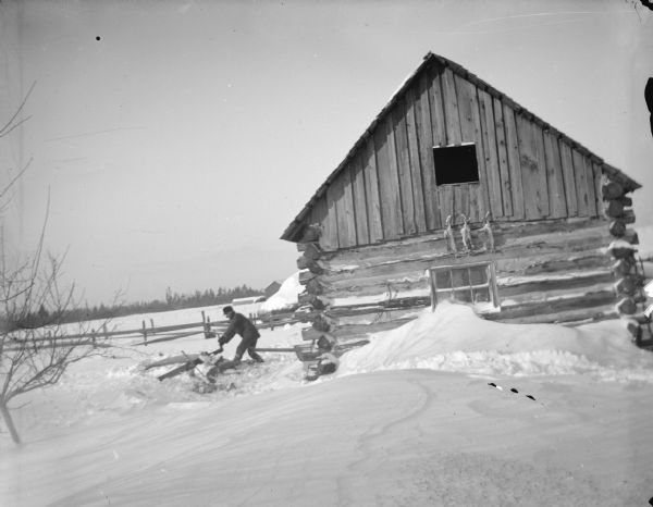 View across deep snow towards a man sawing logs next to a log cabin. In the background is a fence, and beyond across a field are more farm buildings and a line of trees. Above the window on the log cabin are hanging three animal pelts.