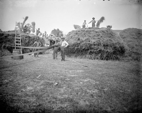 Group portrait of a large group of men working with a threshing machine. In the left foreground is a belt connected to equipment (out of frame) that is running the threshing machine. Men are standing around the threshing machine which is set up between large stacks of hay. Other men are standing on the haystacks ready to load the threshing machine. The roof of a barn is in the far background.