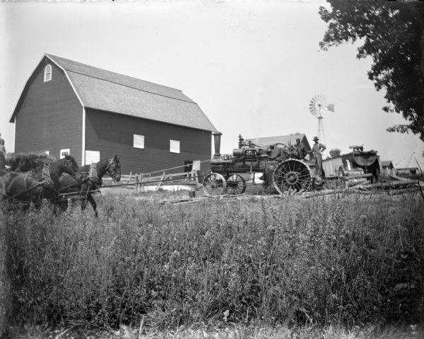 View across field of tall grass towards two men on a steam tractor belt-driving a thresher on the far left near a team of two horses. In the background is are a barn, farm buildings and a windmill.
